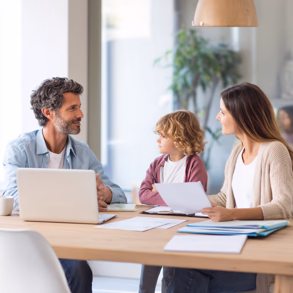 Een man, vrouw en kind zitten aan een tafel met documenten en een laptop, in een lichte kamer met een plant op de achtergrond.