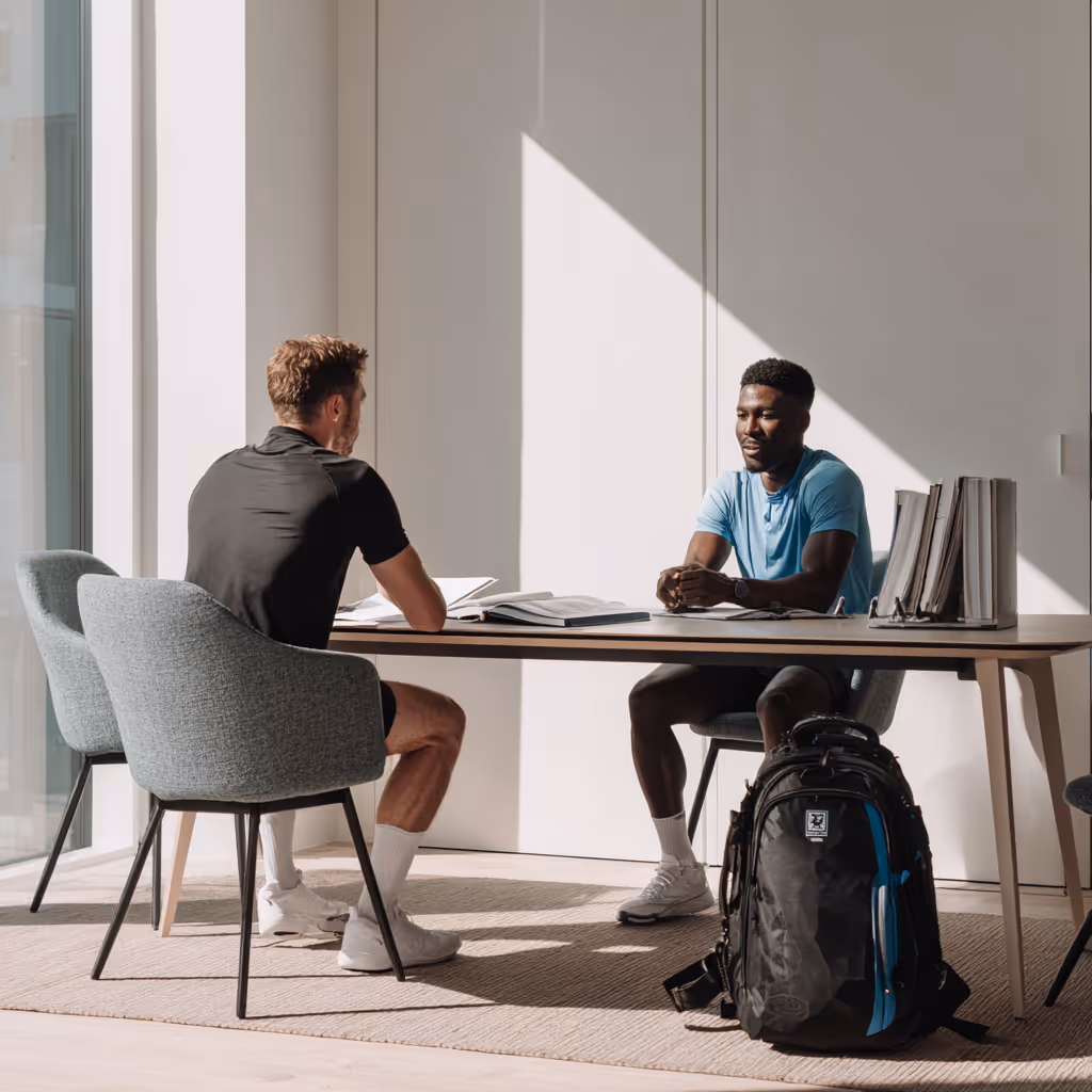 Twee mannen zitten aan een tabel voor een gesprek met boeken en mappen, naast een grote rugzak.