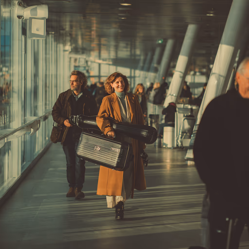 Vrouw in een camelkleurige jas loopt door een lichte luchthaven- of stationsgang met een koffer en een instrumentenkoffer, gevolgd door een man met een zak.