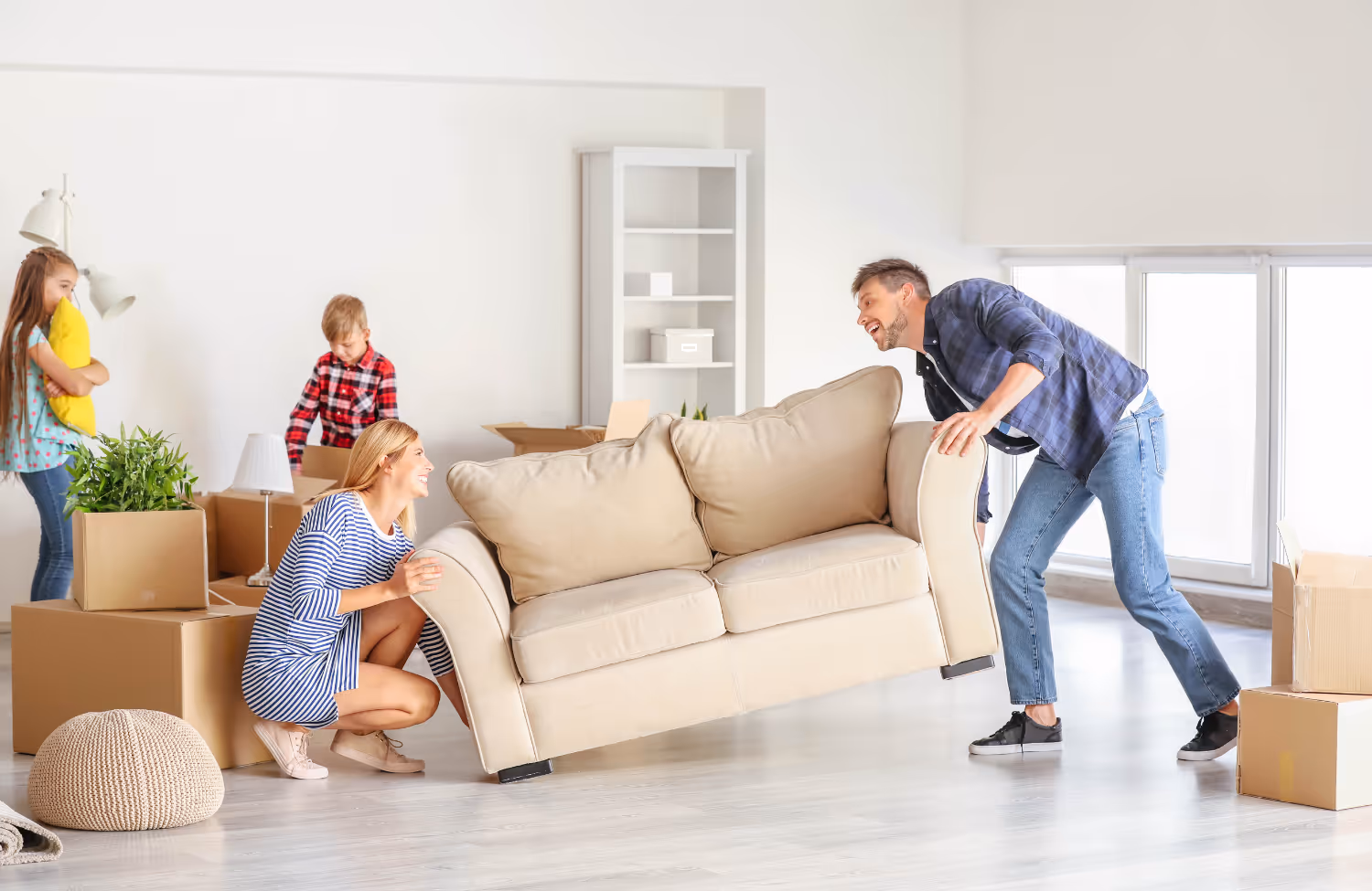 Vader en moeder tillen samen een beige bank in een lege kamer, terwijl twee kinderen dozen uitpakken.