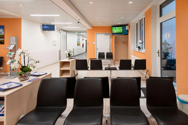 Moderne Wartezimmer mit schwarzen Stühlen, orangefarbenen Wänden und zwei Fernsehern an der Wand.