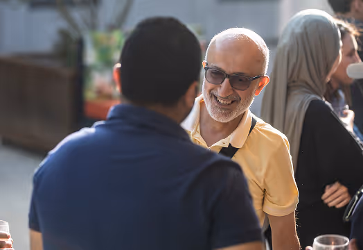 Un homme souriant avec lunettes et polo jaune discute avec un autre homme de dos lors d'une réunion sociale en plein air.