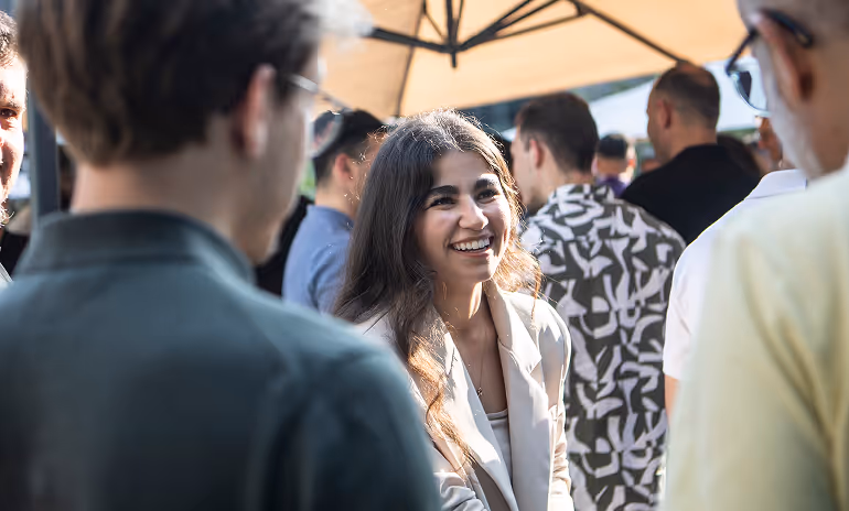 Une femme souriante en veste claire parle à un groupe de personnes lors d'un événement en plein air sous un parasol.