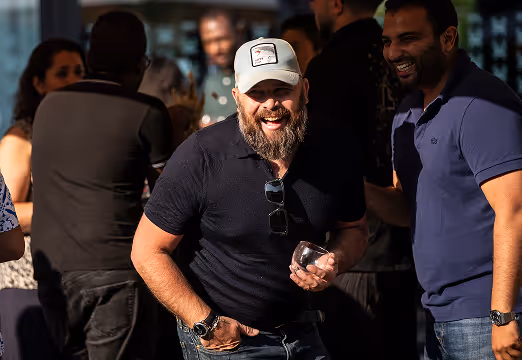 Un homme barbu portant une casquette blanche et un polo noir rit en tenant un verre lors d'une réunion sociale en plein air avec plusieurs personnes en arrière-plan.