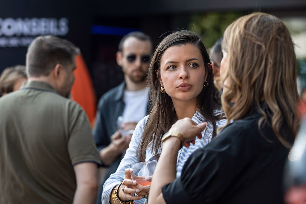 Deux femmes en conversation lors d’un événement social, une tenant un verre, avec d’autres personnes floues en arrière-plan.