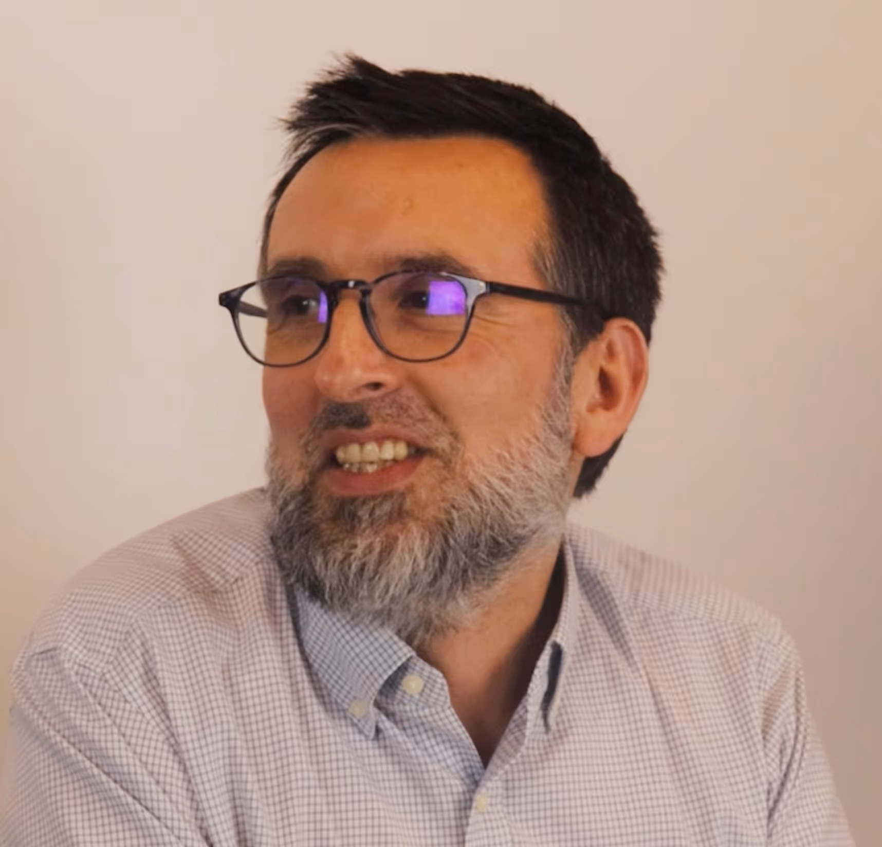 Homme portant des lunettes noires et une chemise à carreaux clairs, souriant et regardant vers la droite.