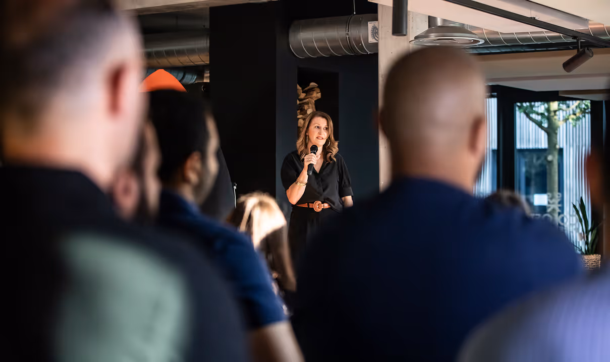 Une femme tenant un micro s'exprimant devant un public, dans un espace intérieur moderne.