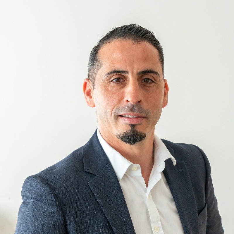 Portrait d'un homme avec un costume bleu marine et une chemise blanche, souriant légèrement devant un fond clair.