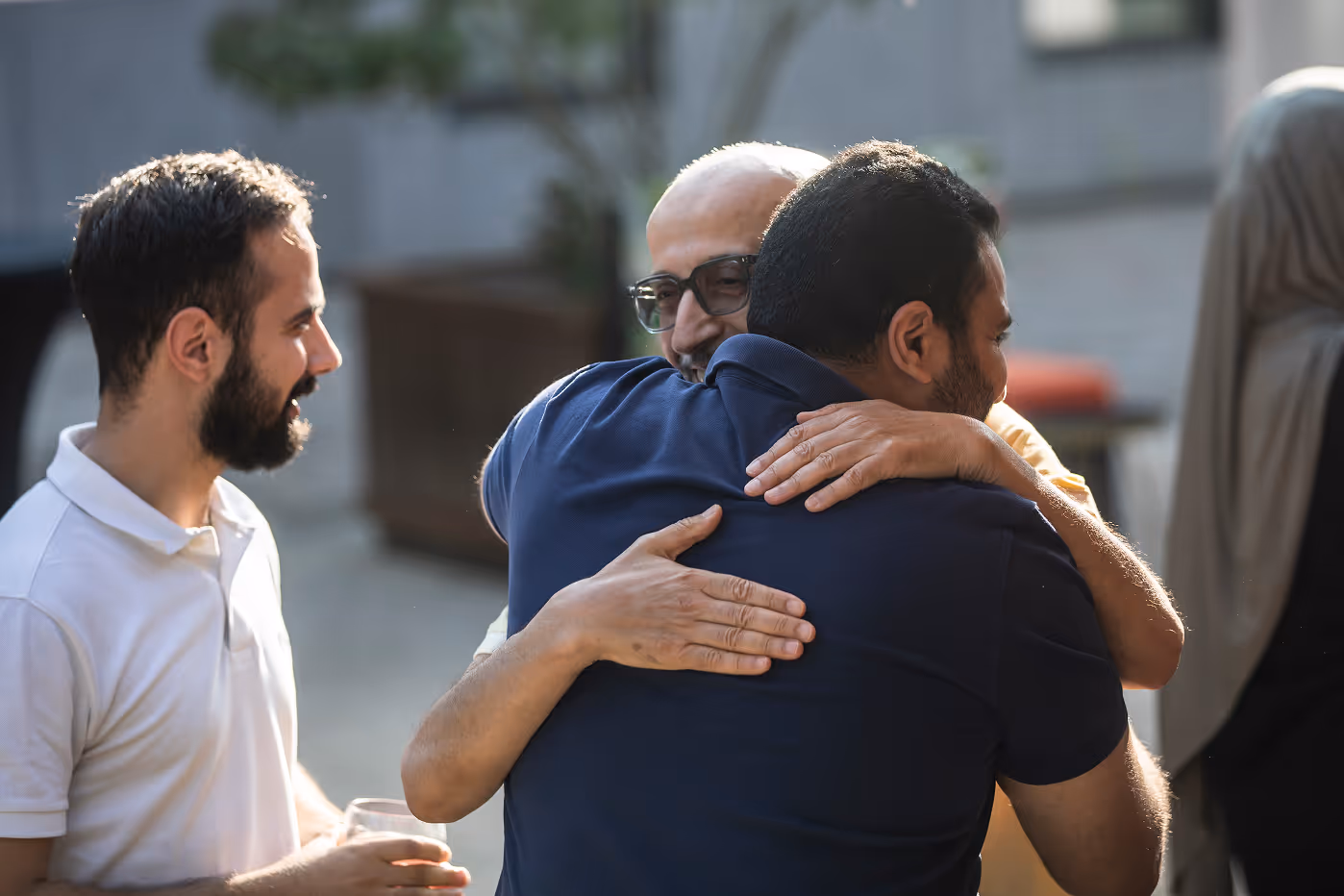 Deux hommes s'embrassant chaleureusement tandis qu'un troisième homme souriant les regarde avec un verre à la main.