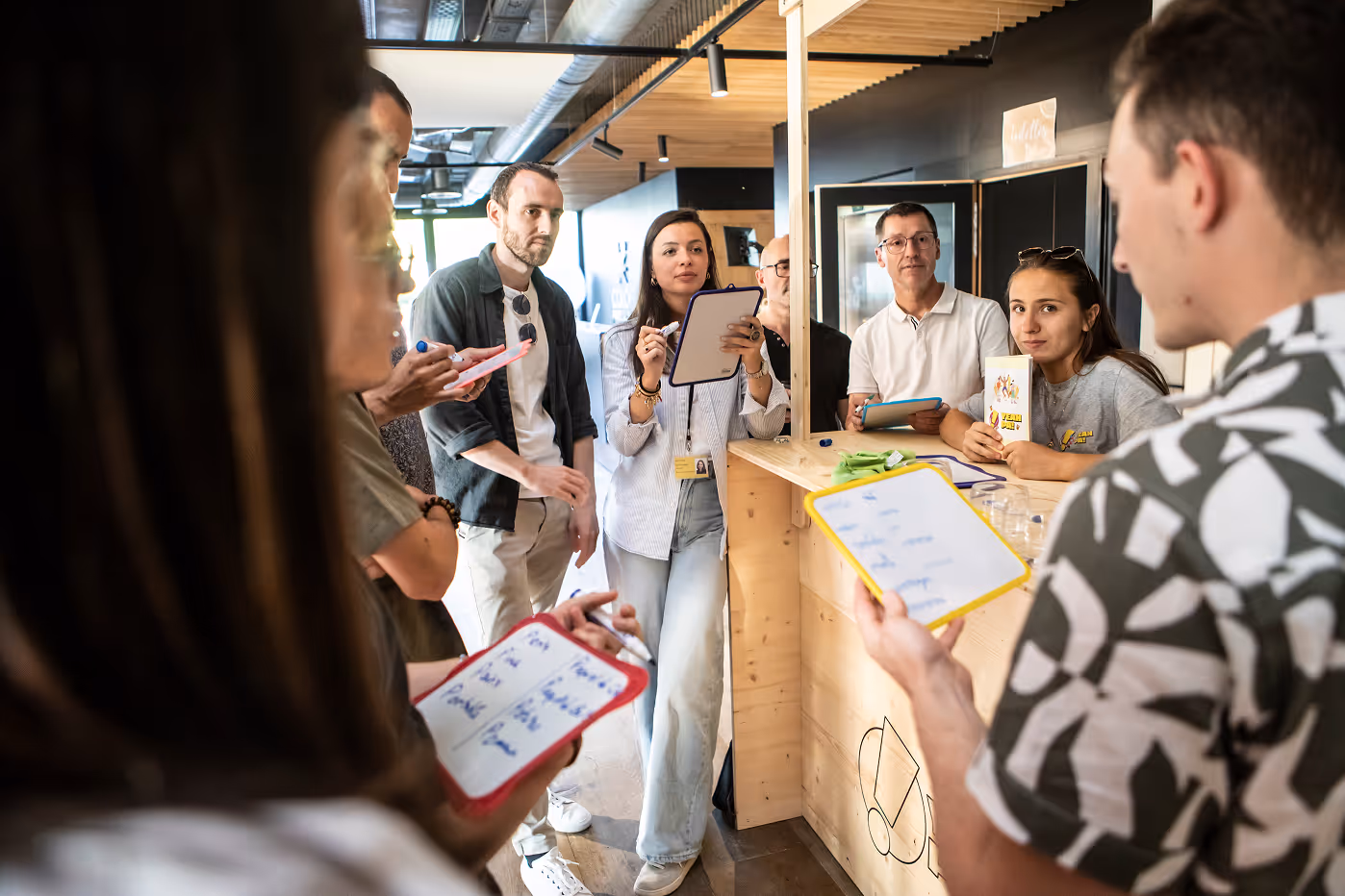 Un groupe de jeunes adultes prend des notes sur des tableaux blancs lors d'une discussion en intérieur autour d'un comptoir en bois.
