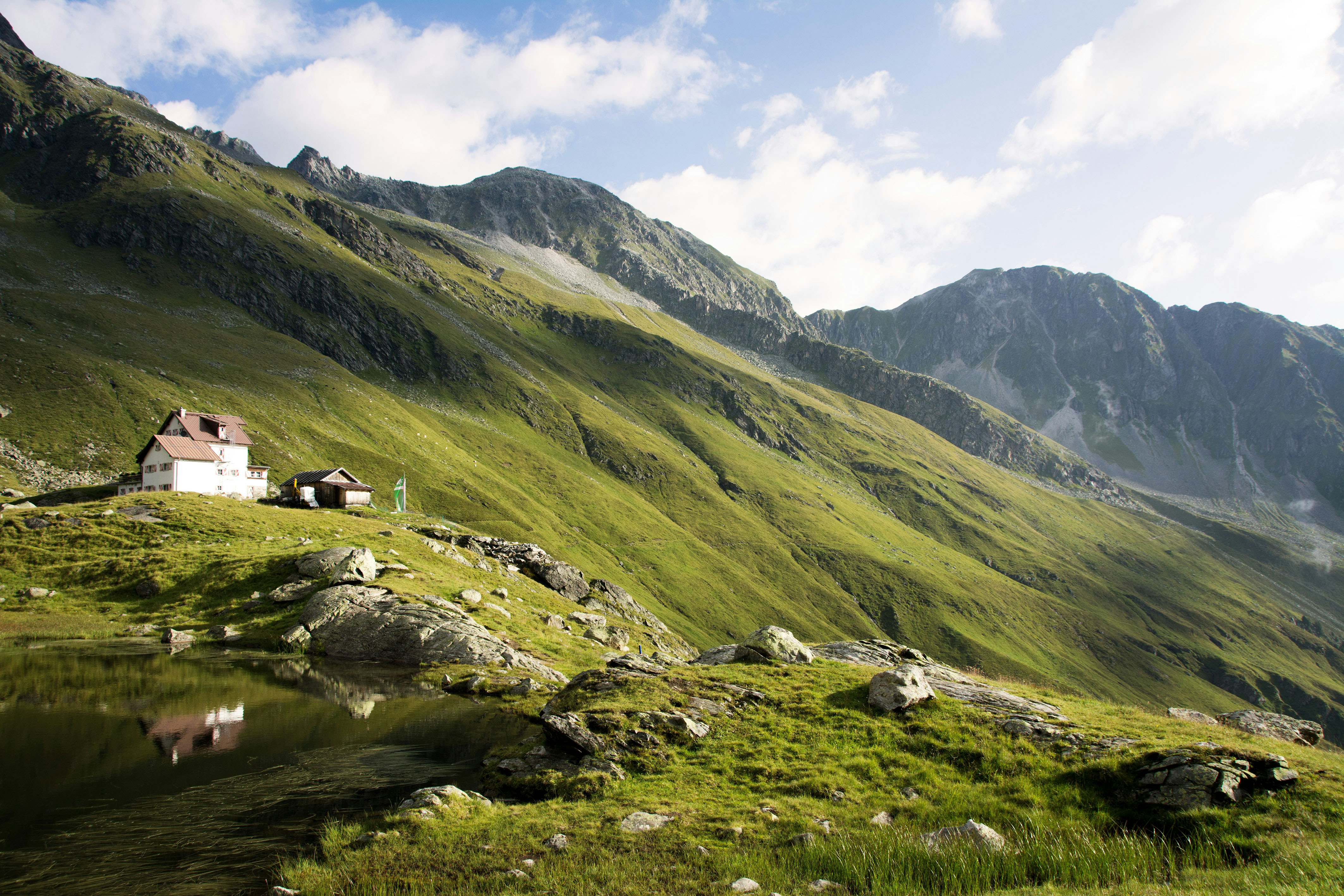 Alpenhütte neben kleinem Teich mit grünen Hügeln und Bergen im Hintergrund unter blauem Himmel.