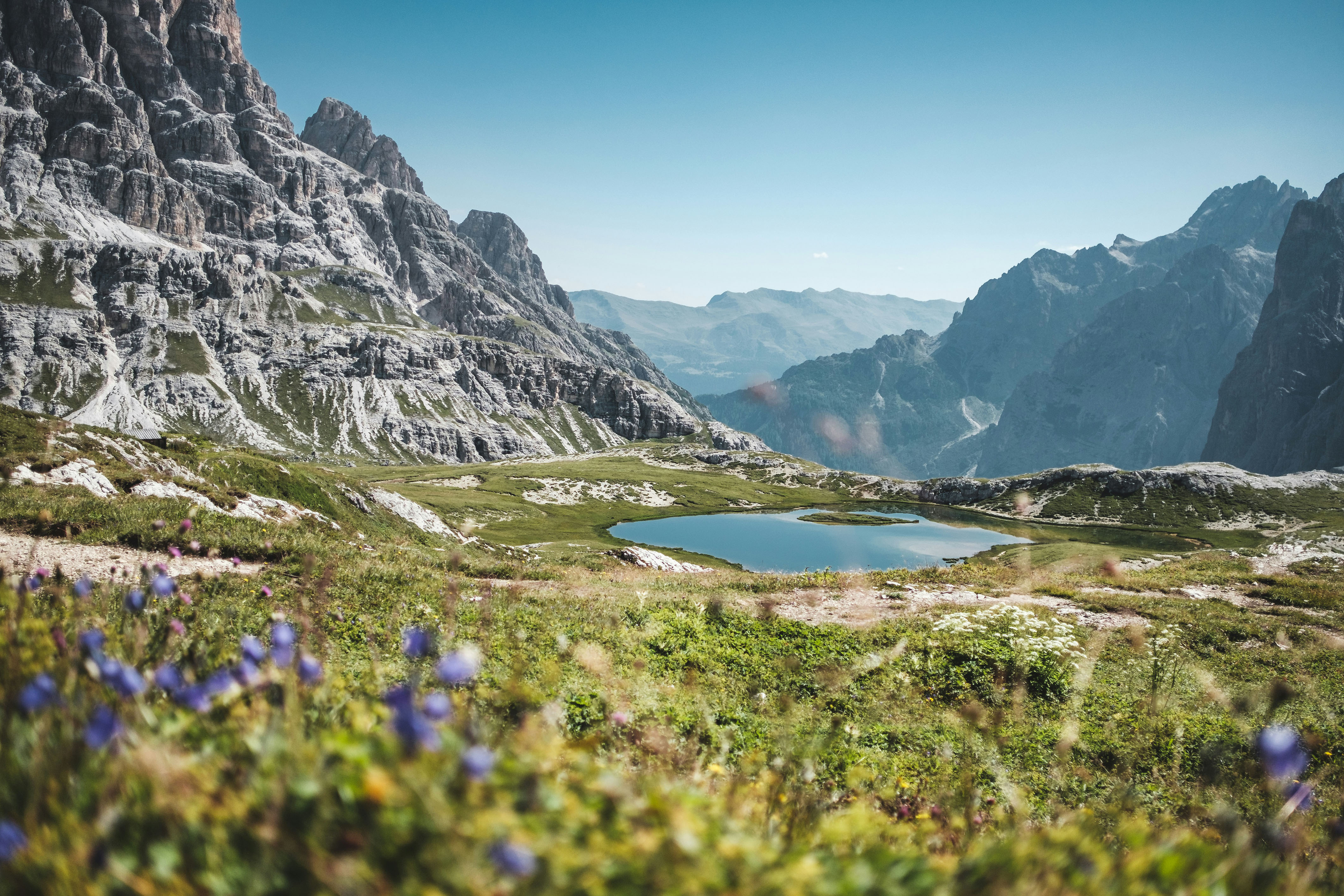 Berglandschaft mit einem klaren Bergsee, grünen Wiesen und wilden Blumen im Vordergrund unter blauem Himmel.