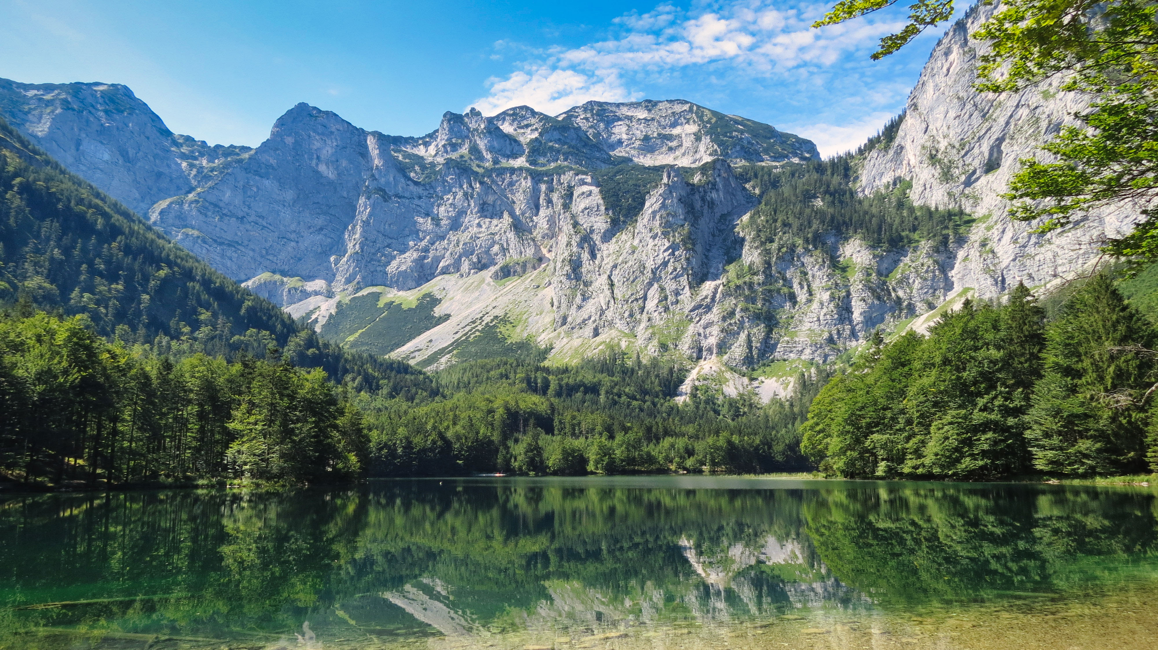 Klarer Bergsee mit Spiegelung von bewaldeten Hängen und steilen Felsen unter blauem Himmel.