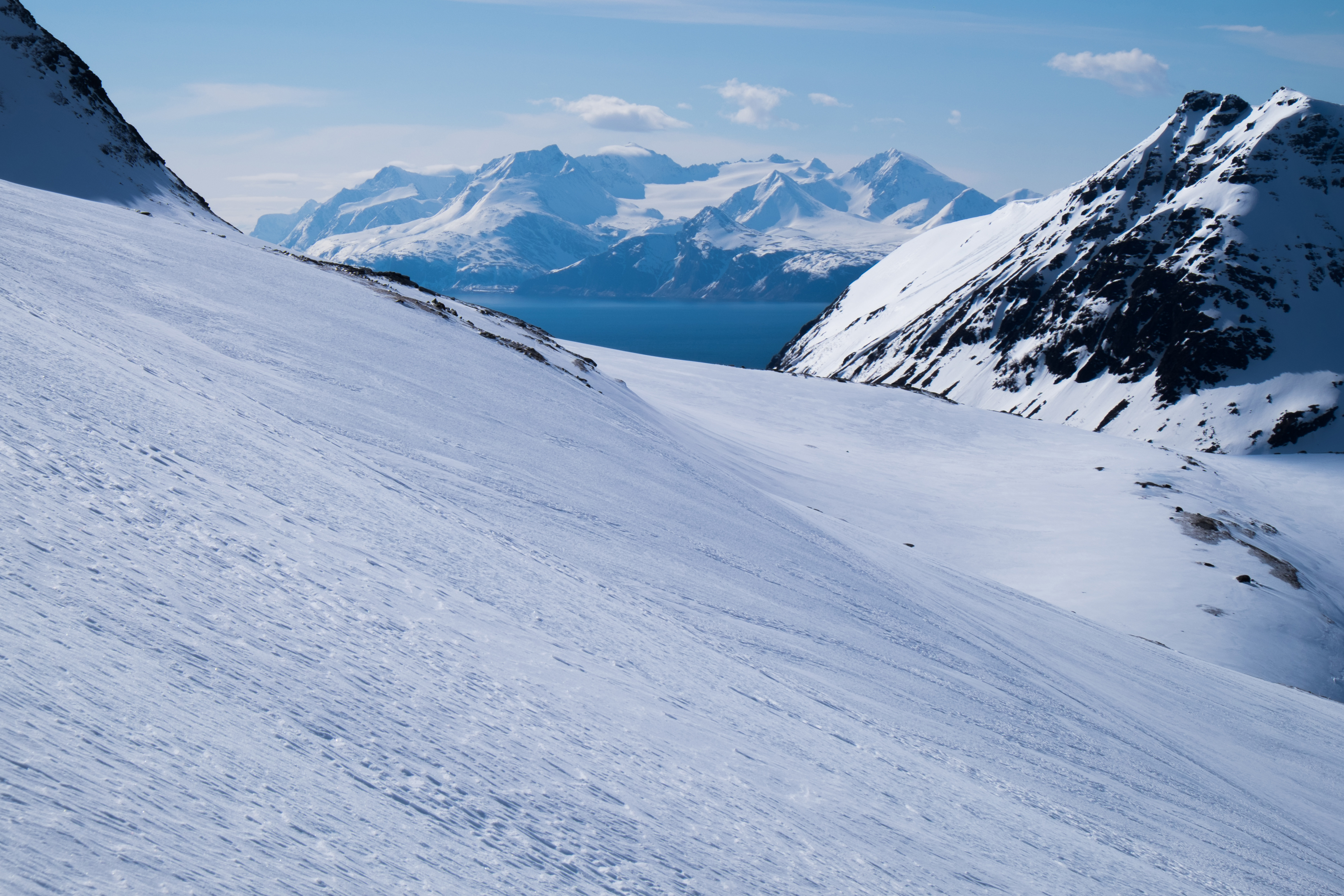 Schneebedeckte Berghänge mit Blick auf schneebedeckte Gipfel und einen See unter blauem Himmel.