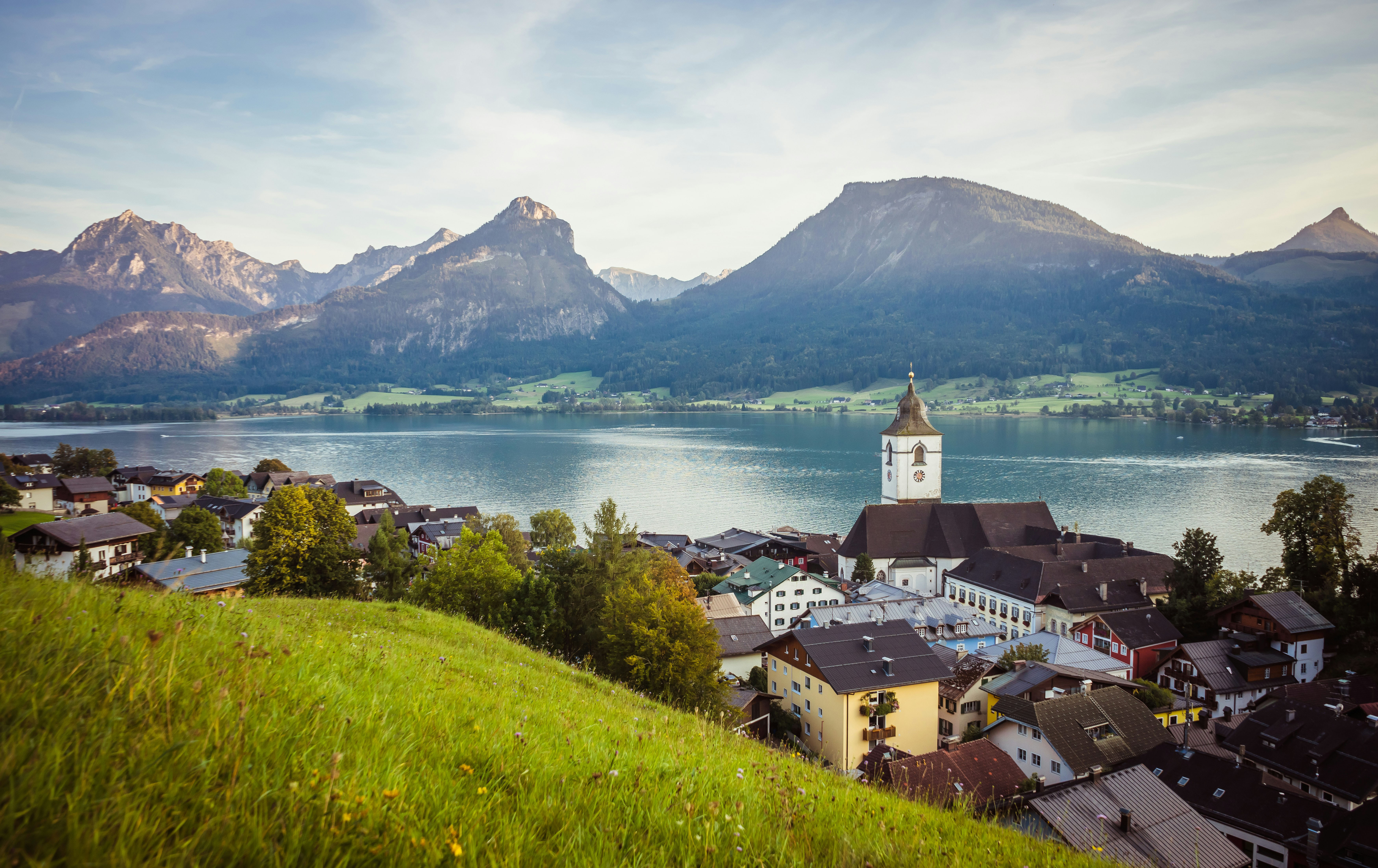 Dorf am See mit Kirche im Vordergrund und Bergen im Hintergrund an einem klaren Tag.