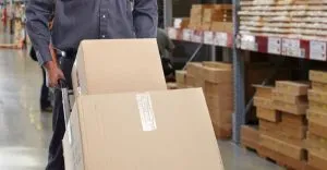 Worker moving large cardboard box in warehouse with stacked wooden pallets