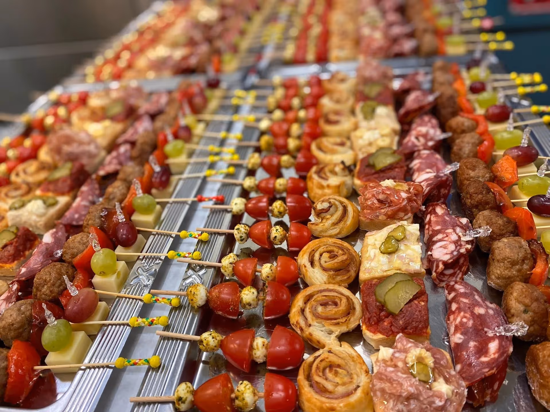 Rows of assorted appetizers including meat skewers, cheese cubes with grapes, tomato and mozzarella skewers, and pastry spirals on a metal tray.