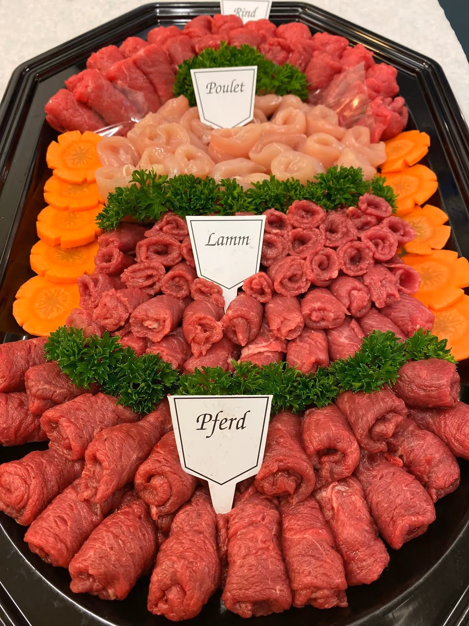 Tray with rows of rolled raw meats labeled Pferd (horse), Lamm (lamb), Poulet (chicken), and Rind (beef), garnished with parsley and sliced carrots.
