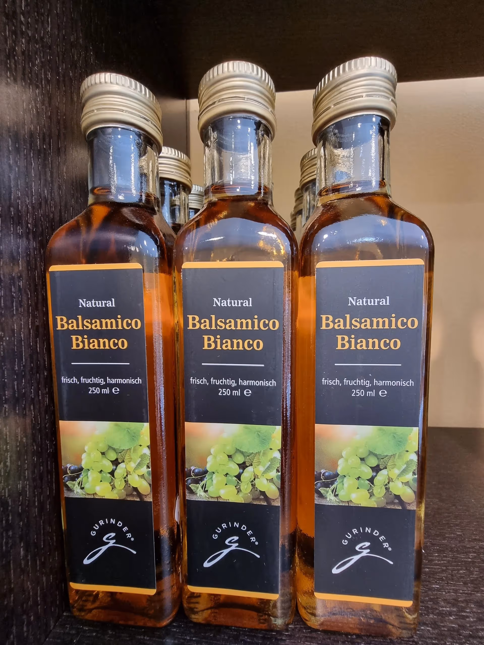 Three bottles of Natural Balsamico Bianco vinegar with beige caps and labels featuring green grapes, displayed on a dark wooden shelf.