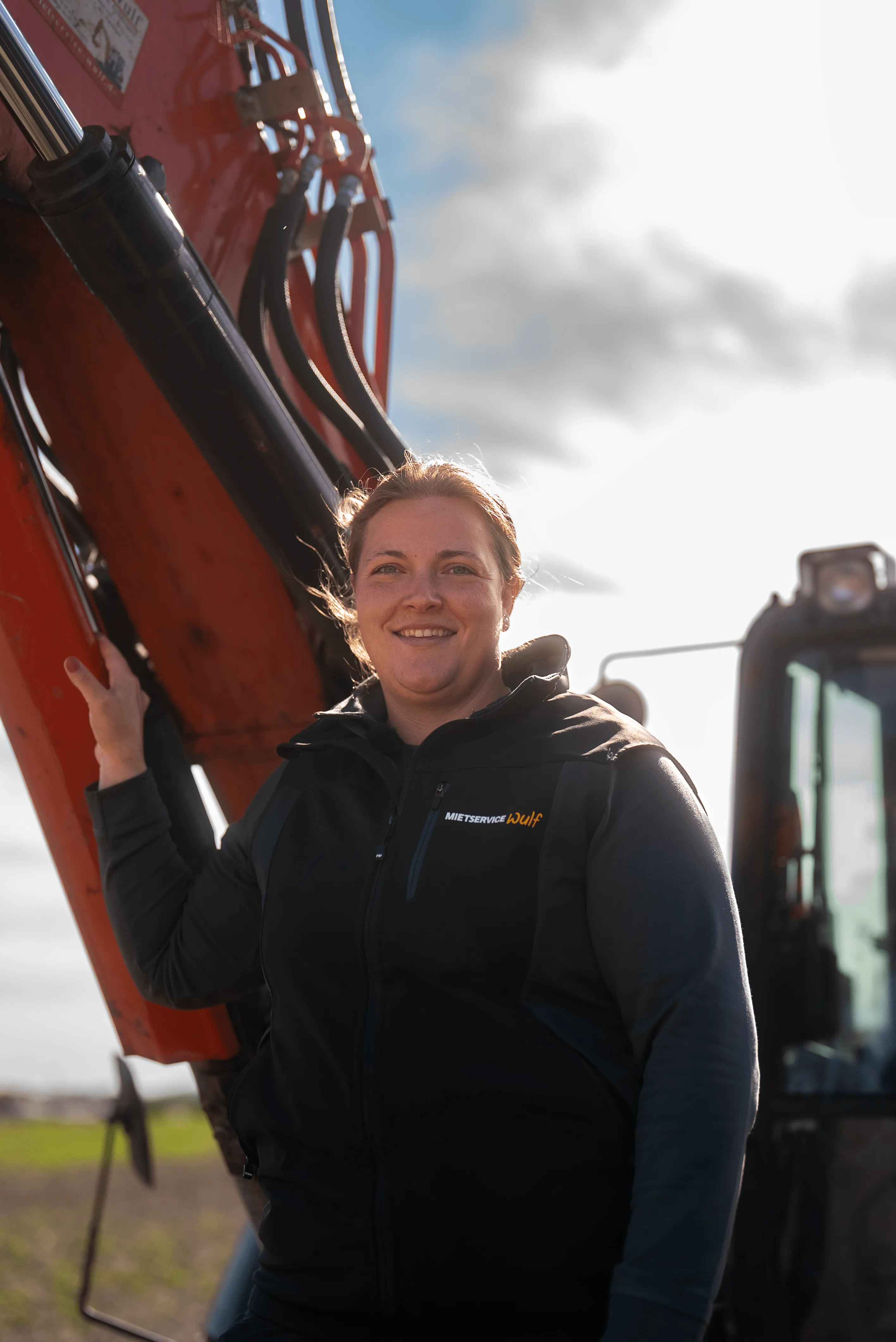 Frau in schwarzer Jacke mit der Aufschrift „Mietservice Wulf“ lächelt, lehnt an einem roten Hydraulikarm einer Baumaschine bei bewölktem Himmel.