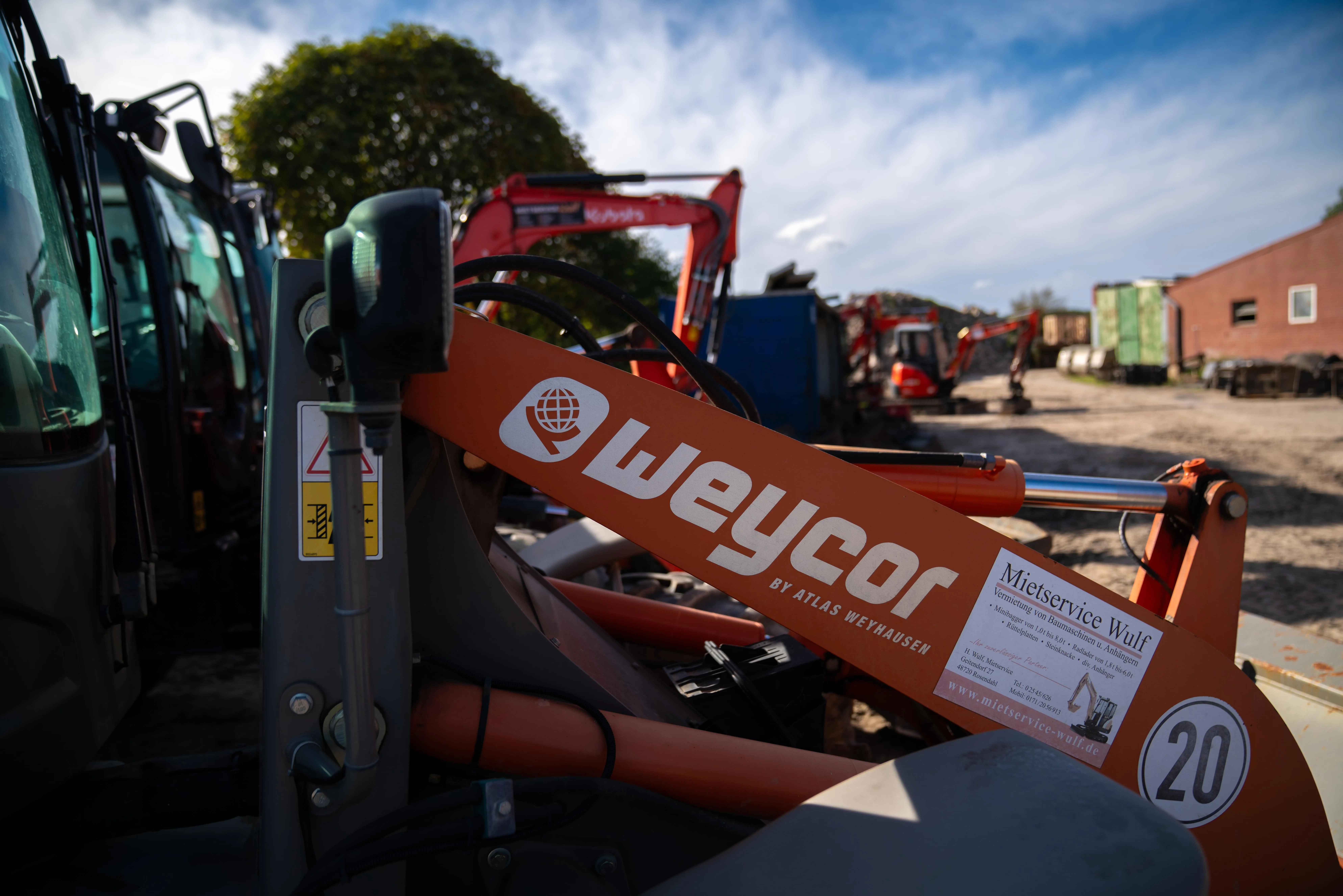 Nahaufnahme eines orangefarbenen Weycor-Baggers auf einer Baustelle mit weiteren Baumaschinen und einem blauen Himmel im Hintergrund.
