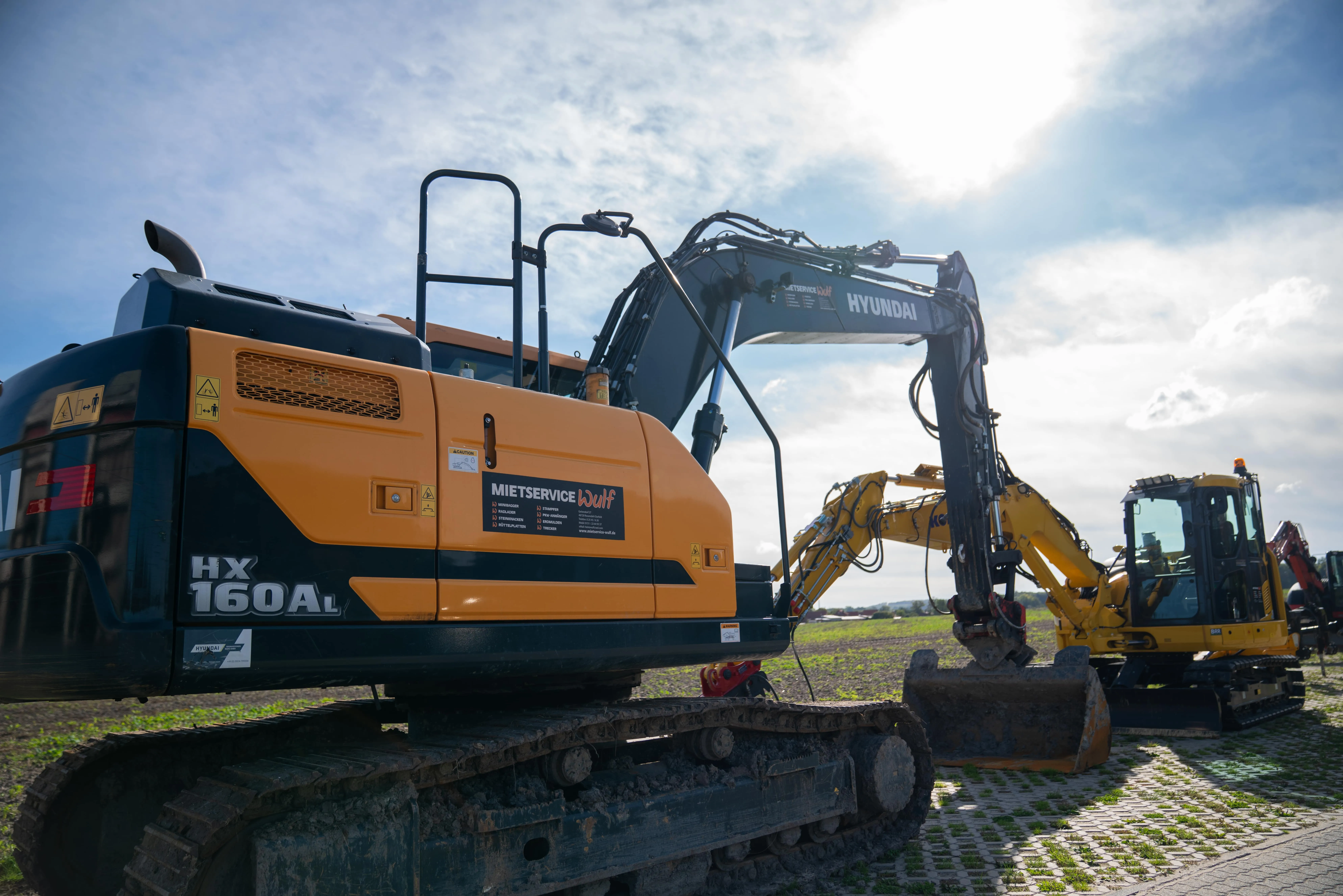 Bagger vom Typ Hyundai HX 160AL und anderer gelber Bagger auf einem befestigten Feld unter bewölktem Himmel.