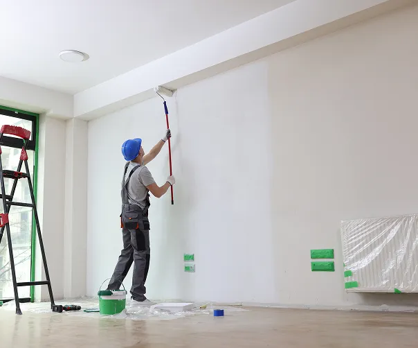 Maler streicht eine Wand mit Farbrolle in einem hellen Raum, Leiter und Farbeimer stehen daneben.