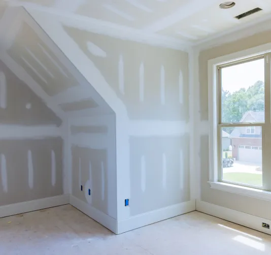 Unfertigter Innenraum mit verputzten Trockenbauwänden und einem Fenster, durch das Tageslicht einfällt.