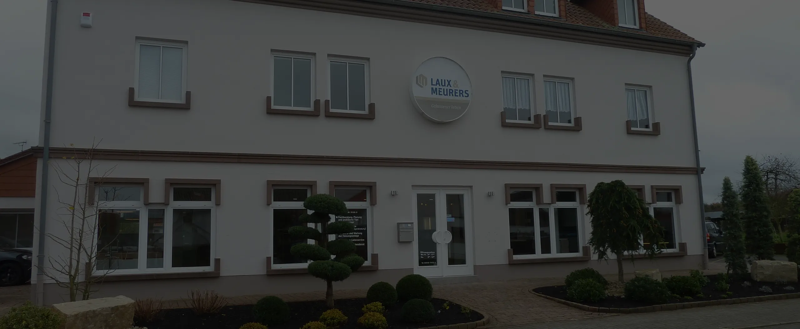 Die Vorderseite eines weißen Bürogebäudes mit mehreren Fenstern, einer Doppeltür und einem rundem Schild mit der Aufschrift ‚LAUX & MEURERS‘, umgeben von gepflegten Pflanzen und Büschen.
