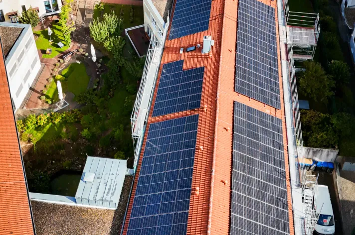Vogelperspektive auf ein Gebäude mit einem roten Ziegeldach, teilweise bedeckt mit Solarpaneelen, neben einem grünen Innenhof.