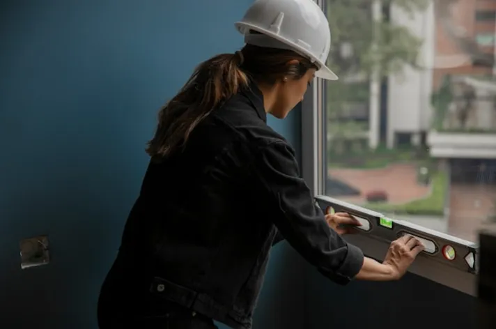 Frau mit Schutzhelm, die mit einer Wasserwaage die Ausrichtung eines Fensters überprüft.