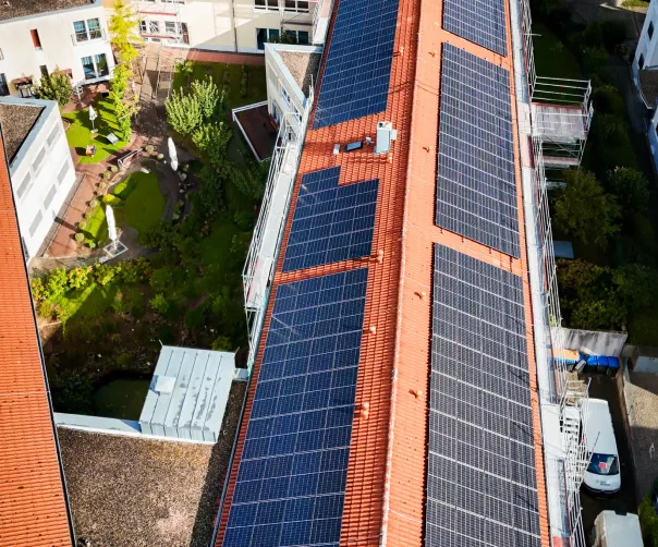 Luftaufnahme eines Gebäudes mit einer Dachfläche, die vollständig mit Solarpanelen bedeckt ist, umgeben von Grünflächen und anderen Gebäuden.