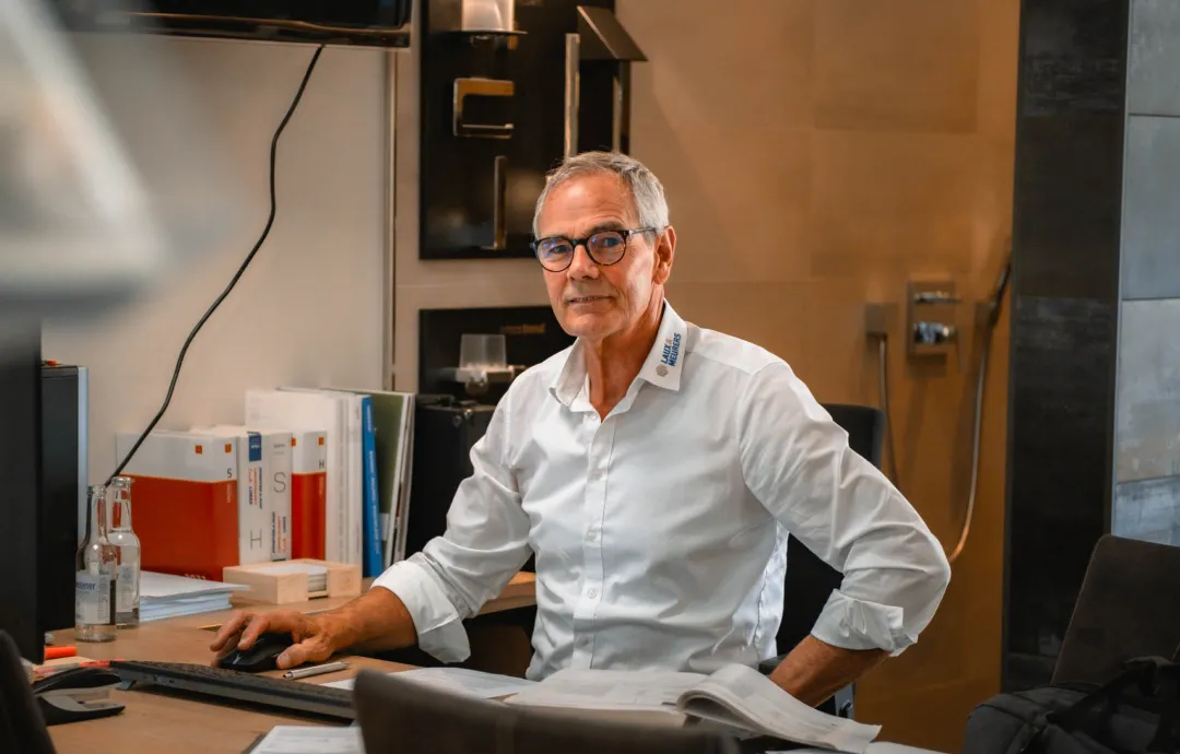 Älterer Mann mit Brille sitzt an einem Schreibtisch mit geöffnetem Buch und Computertastatur.