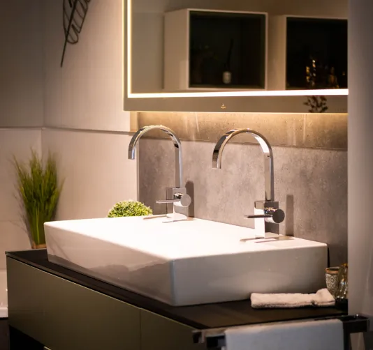 Modernes Badezimmer mit weißem Doppelwaschbecken, zwei verchromten Wasserhähnen, Spiegel mit Beleuchtung und Pflanzen