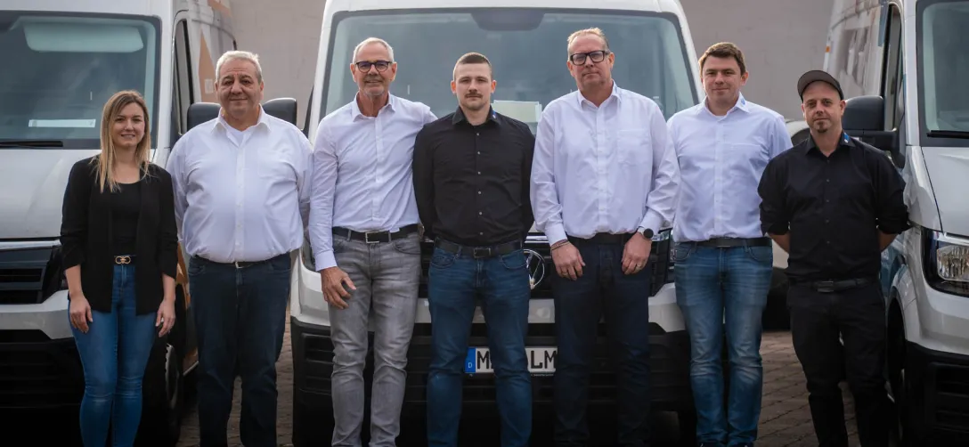 Siebzehn Mitarbeiter stehen vor weißen Transportern, einige tragen weiße Hemden, andere schwarze Hemden oder schwarze Jacken.