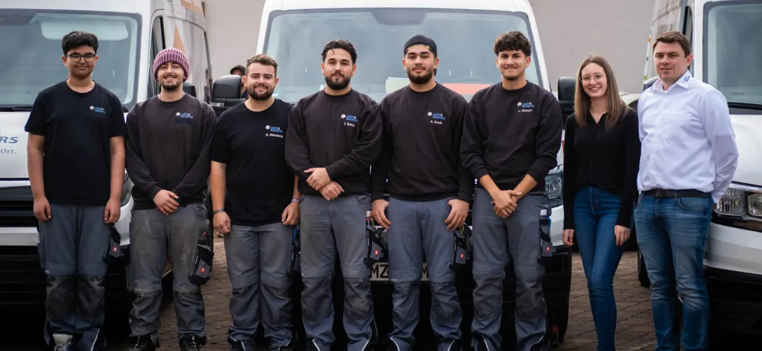 Acht Personen stehen in einer Reihe vor weißen Transportern, sieben Männer tragen Arbeitskleidung, eine Frau und ein Mann links außen tragen Alltagskleidung.