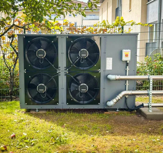 Außeneinheit einer Wärmepumpe mit vier großen schwarzen Lüftern, installiert auf einer Rasenfläche neben einem Gebäude.