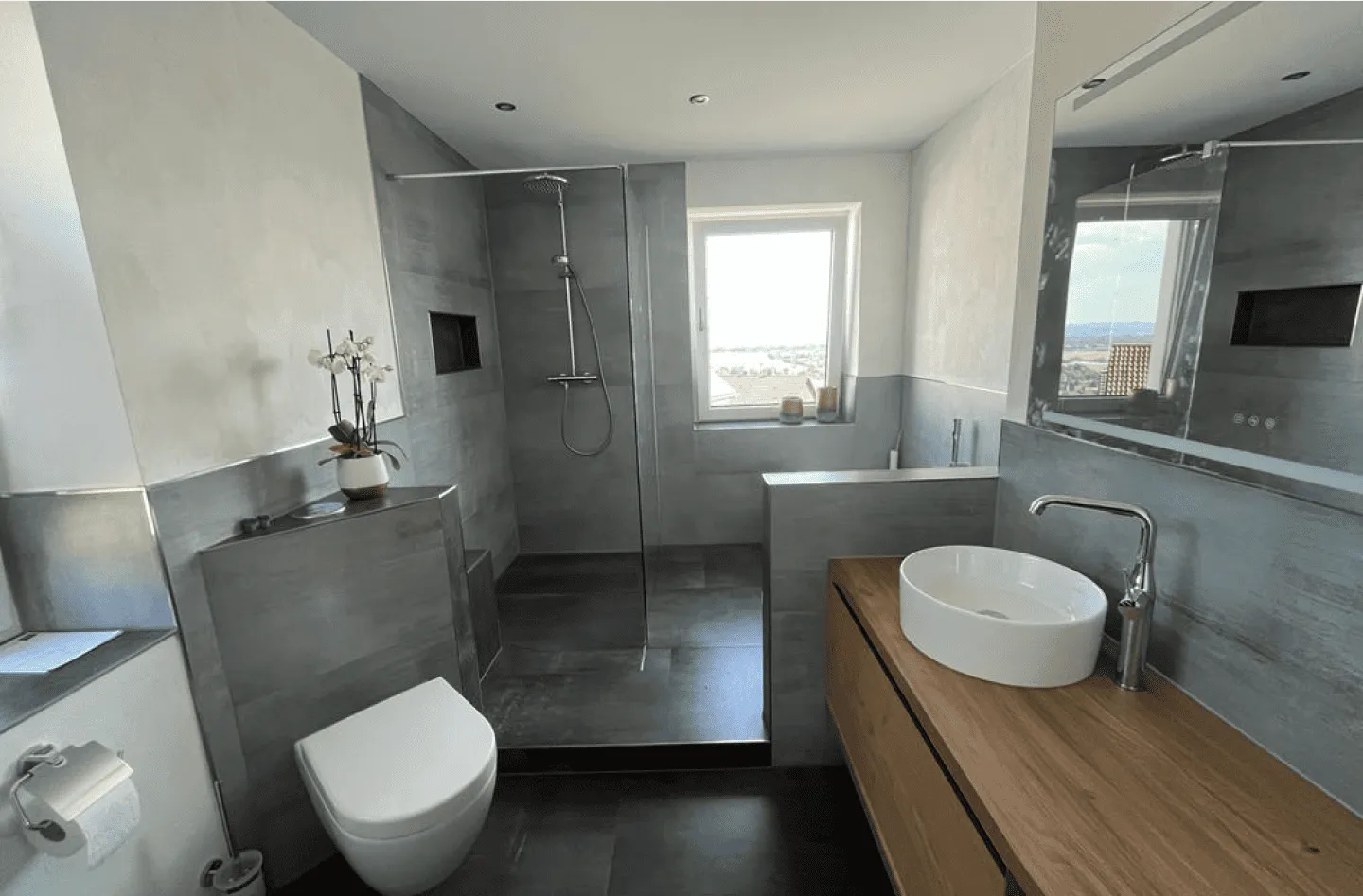 Modernes Badezimmer mit Glasdusche, weißer Toilette, rundem Aufsatzwaschbecken auf Holzunterschrank und Fenster mit Blick nach draußen.