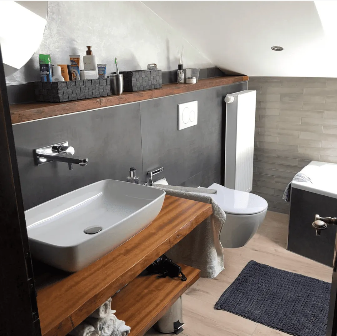 Modernes Badezimmer mit weißem Waschbecken auf Holzarbeitsplatte, wandhängendem WC, grauer Fliesenwand und Badewanne unter einer Schräge.