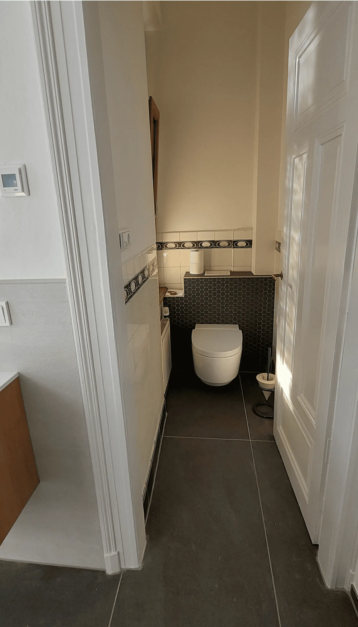 Blick in eine kleine Toilette mit weißer Wandtoilette, dunkelgrauen Bodenfliesen und weißen Wandfliesen mit schwarzem Musterband.