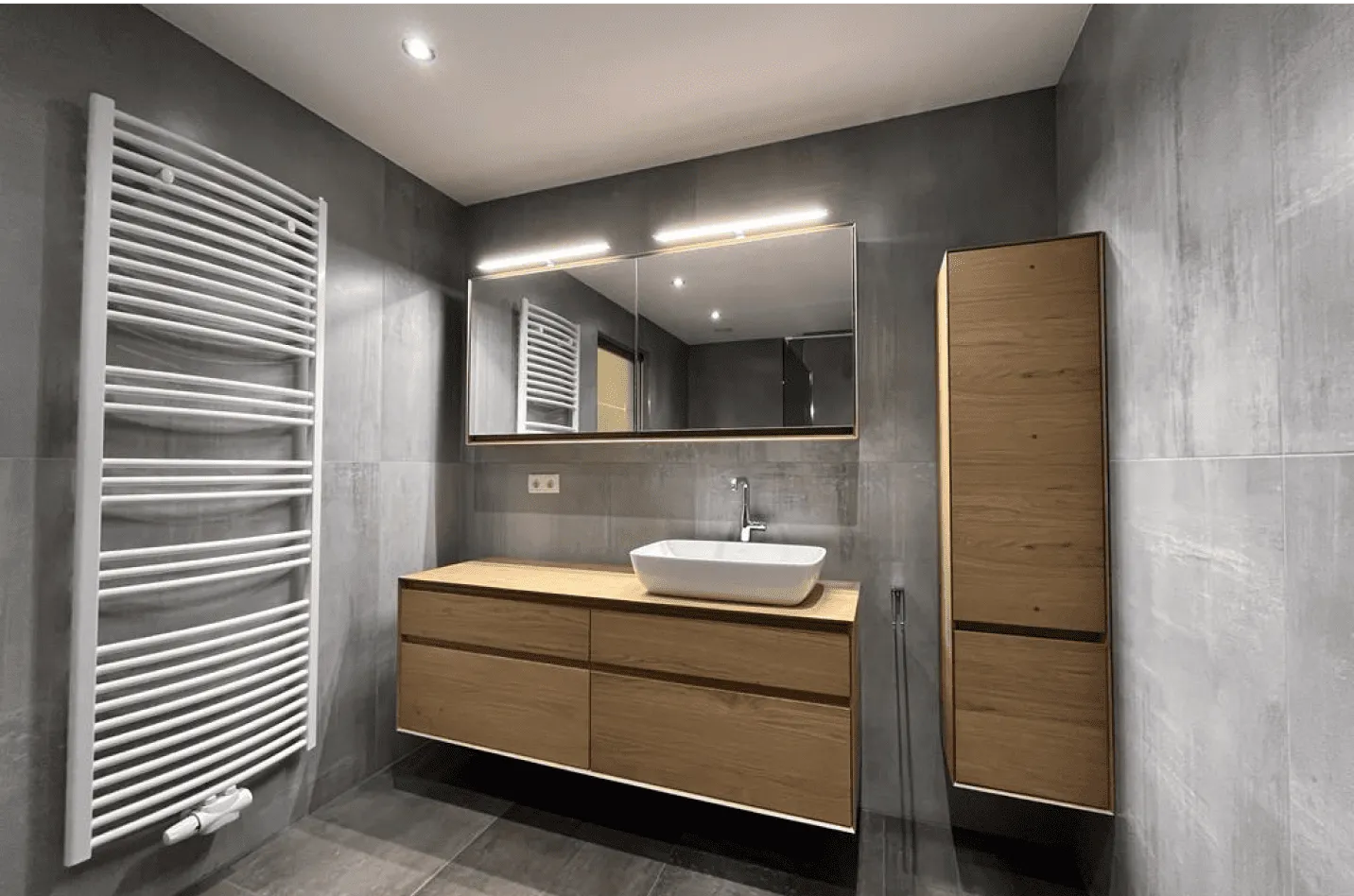 Modernes Badezimmer mit grauen Fliesen, hängendem Holzwaschtisch mit weißem Aufsatzbecken, beleuchtetem Spiegel und weißem Handtuchwärmer.