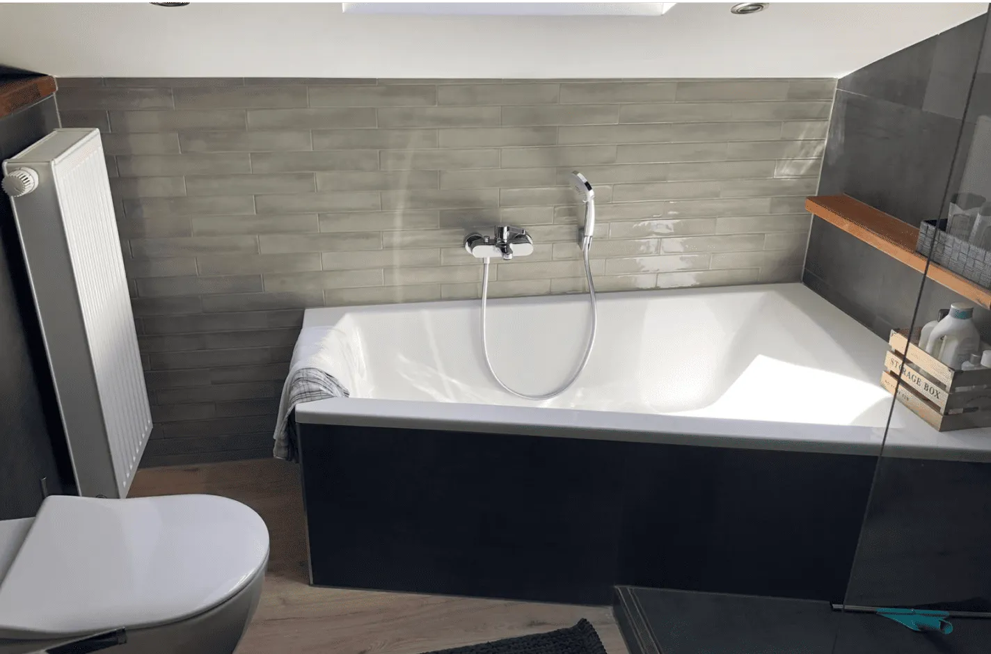 Modernes Badezimmer mit grauer Kachelwand, weißer Badewanne mit Handbrause und einem Handtuch, Toilette, Heizkörper und einem Holzboden.