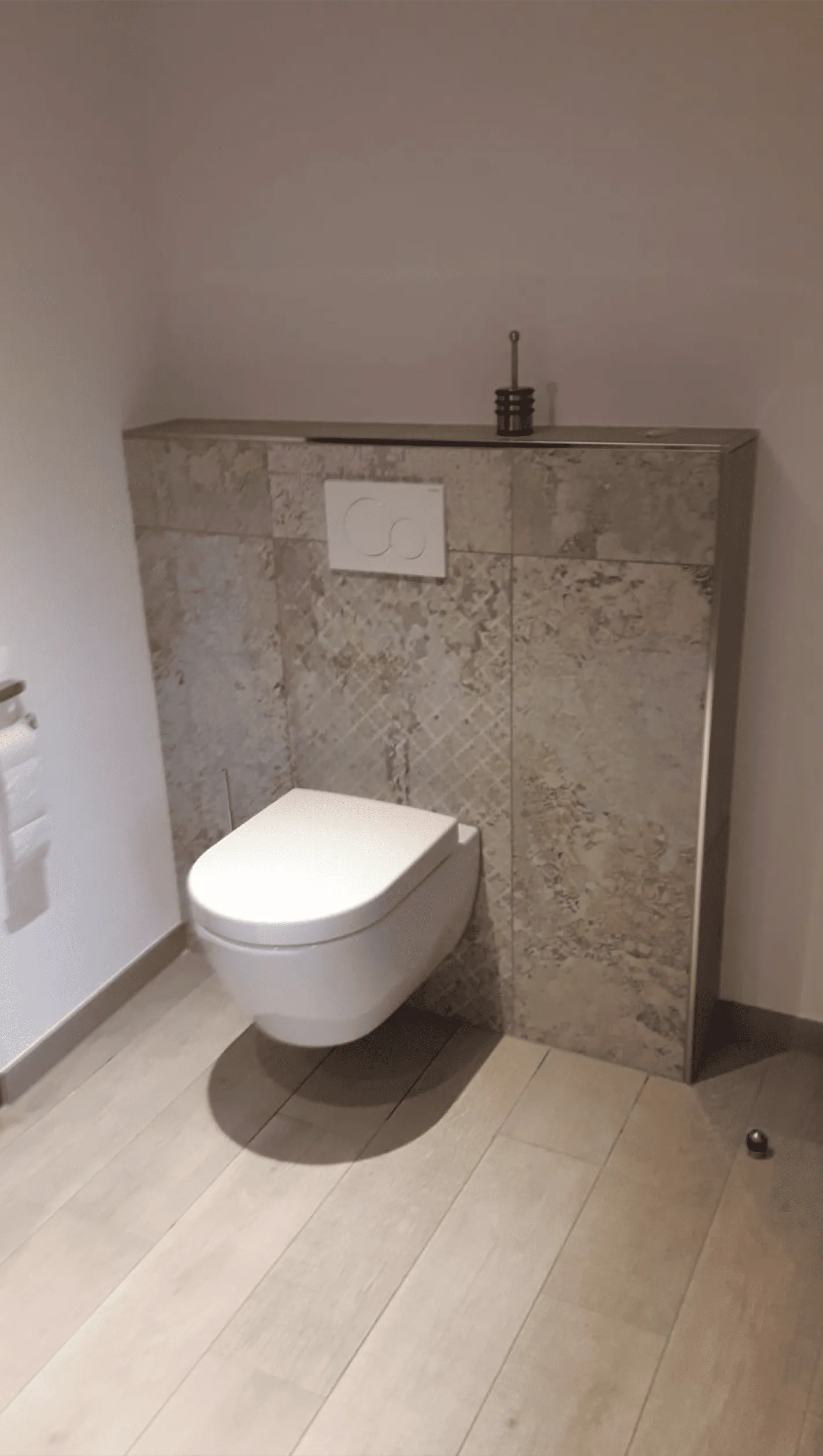 Moderne Wandtoilette mit Befestigung in einer Badezimmerwand mit beige gemusterten Fliesen und Holzoptik-Bodenfliesen.