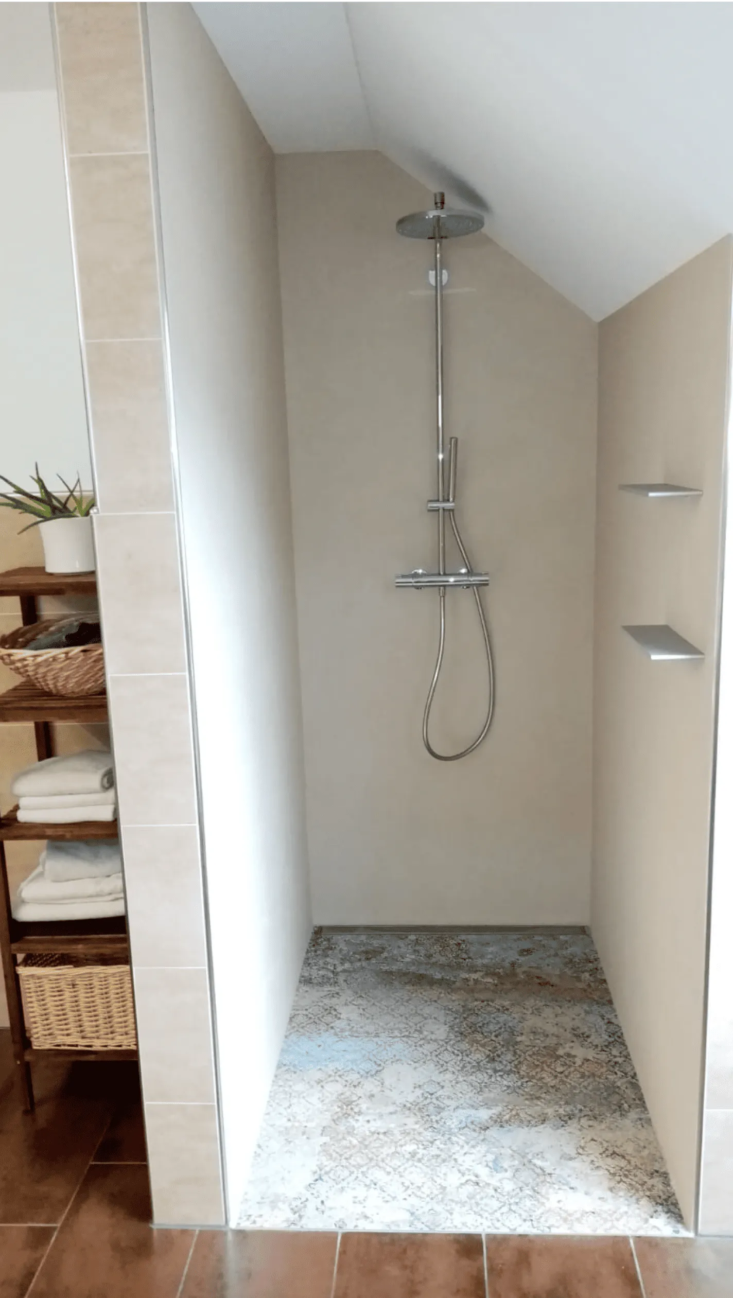 Minimalistische Duschkabine mit regenduschkopf, gealtertem gemustertem Boden und zwei Wandregalen, links ein Holzregal mit gefalteten Handtüchern und einem Pflanzenkübel.