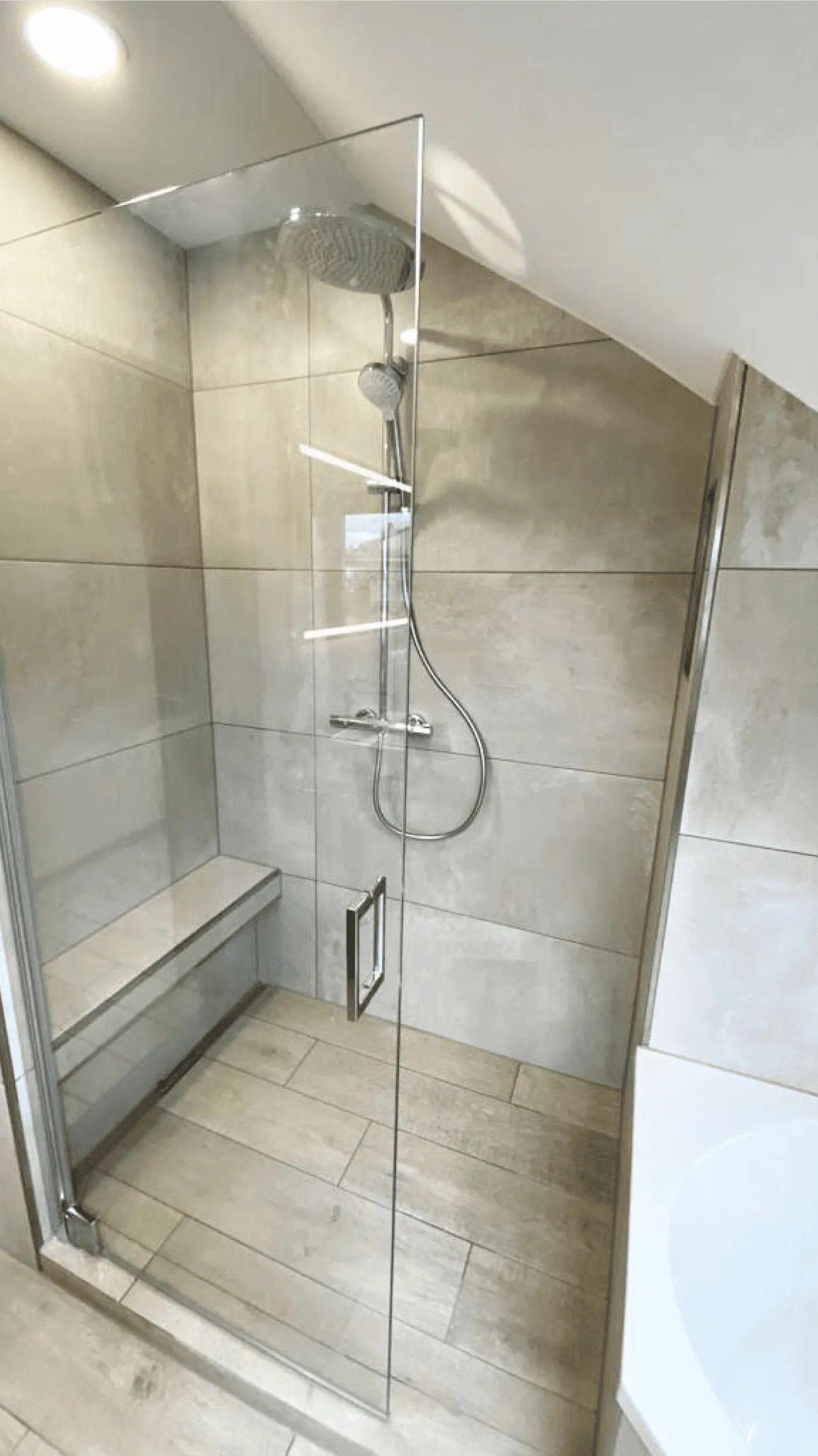 Moderne Dusche mit Glasabtrennung, Regendusche, Handbrause und eingebauter Sitzbank in grauen Fliesen.