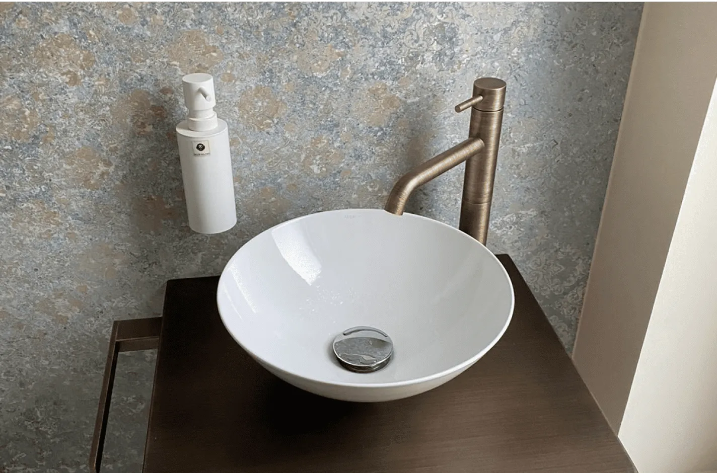 Weißes rundes Waschbecken mit einem kupferfarbenen Wasserhahn und einem weißen Seifenspender an einer gemusterten grauen Wand.