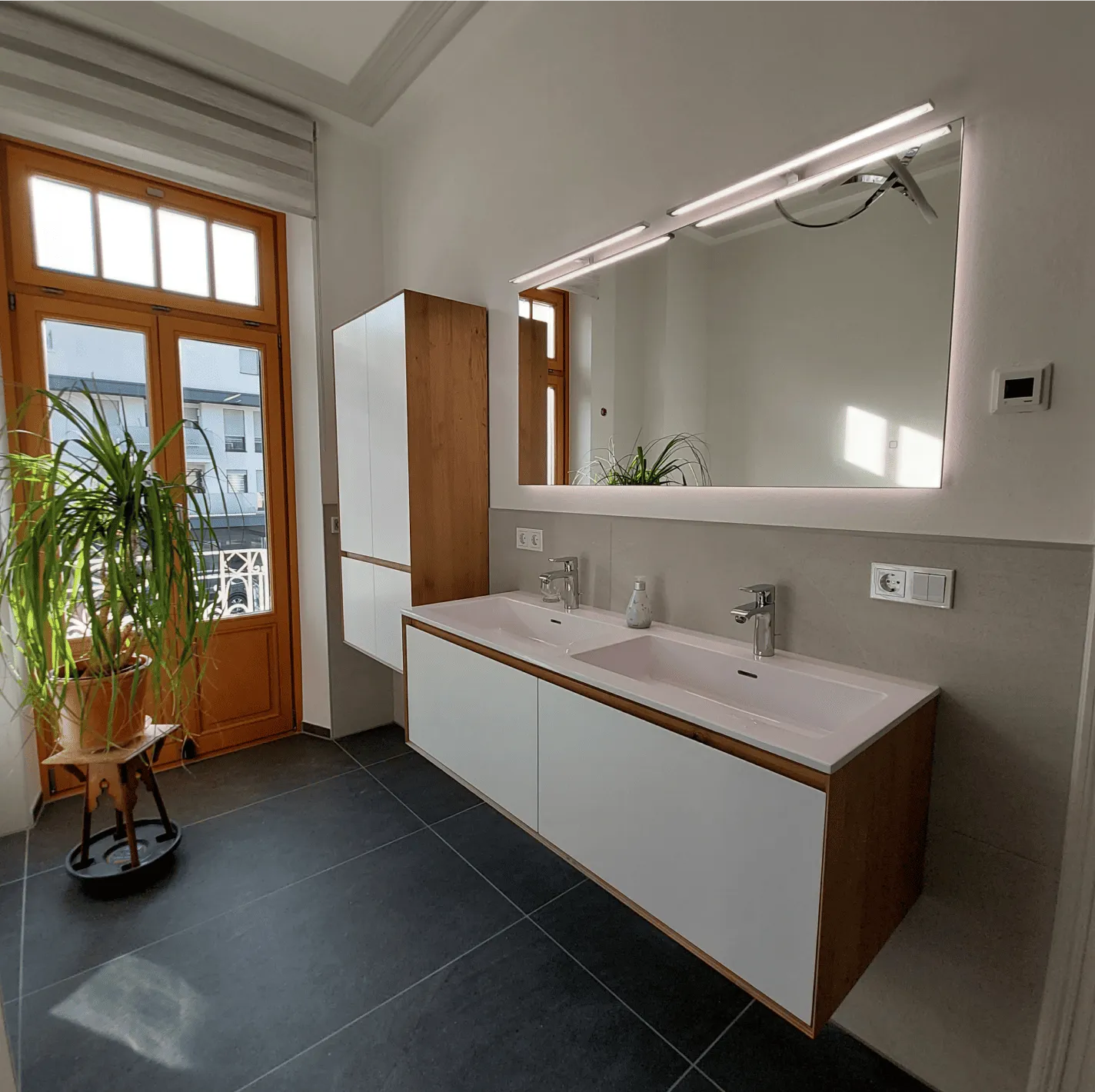 Modernes Badezimmer mit Doppelwaschbecken, großem beleuchtetem Spiegel und Holz-Akzenten neben einer Tür mit großem Fenster und einer Pflanze.