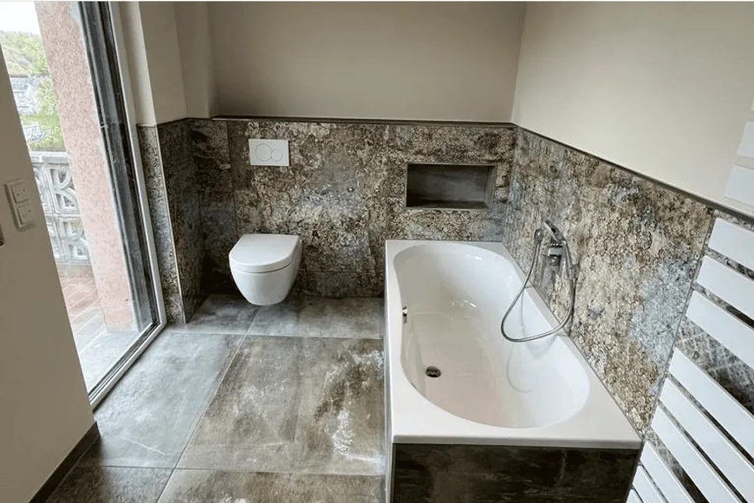 Modernes Badezimmer mit einer weißen Badewanne, wandhängendem WC und grauen Steinfliesen an Boden und Wänden.