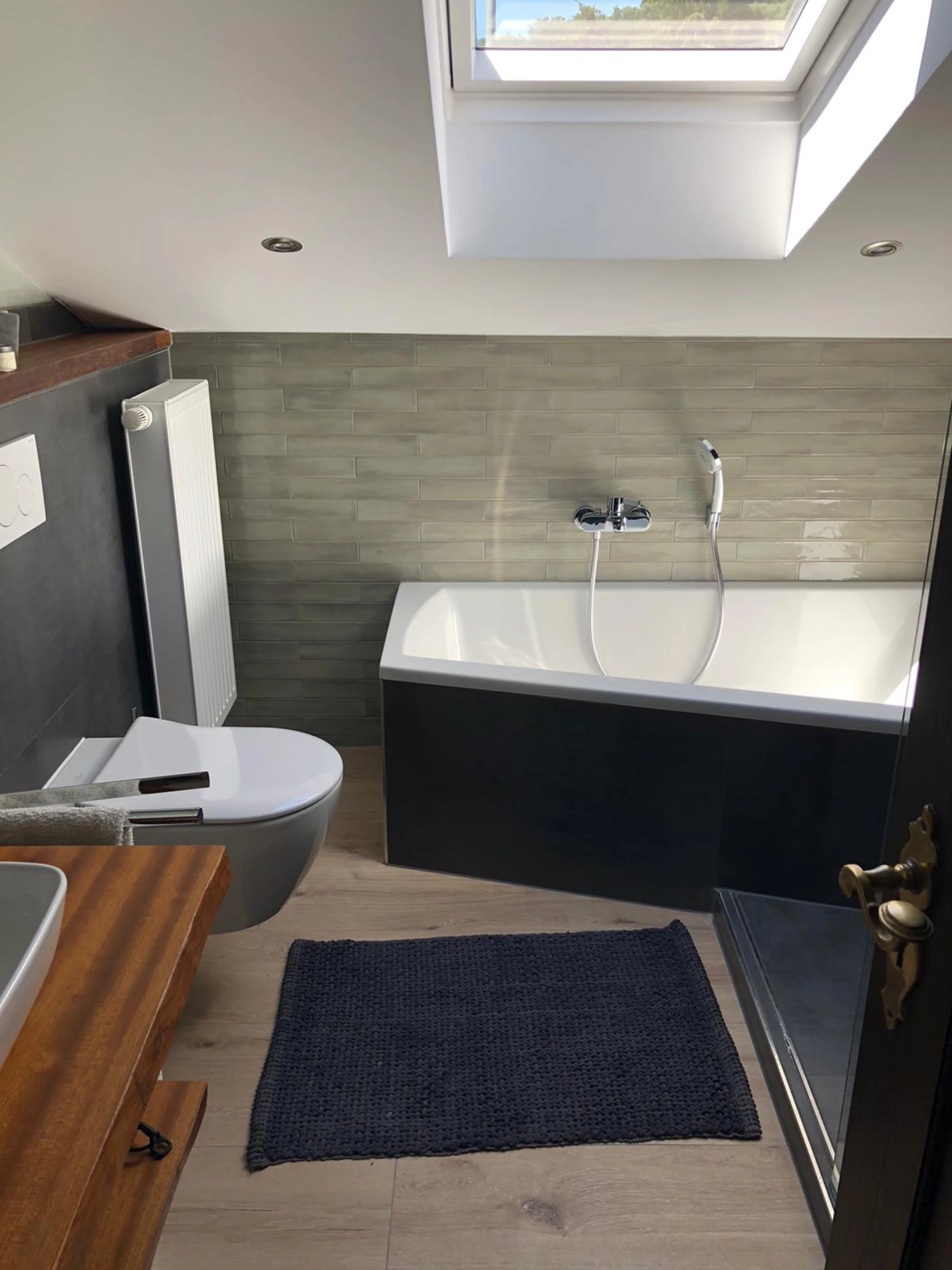 Modernes Badezimmer mit einer weißen Badewanne, grauen Fliesen, einem Holzwaschbecken und einem Dachfenster.