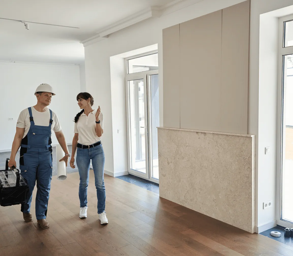 Ein Bauarbeiter mit Helm und Werkzeugkasten und eine Frau im weißen Polo unterhalten sich in einem hellen, modernen, unfertigen Raum mit Holzboden und großen Fenstern.